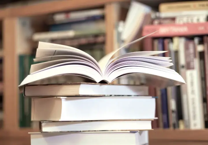 Otwarta książka na stosie innych książek, na tle regału z książkami.